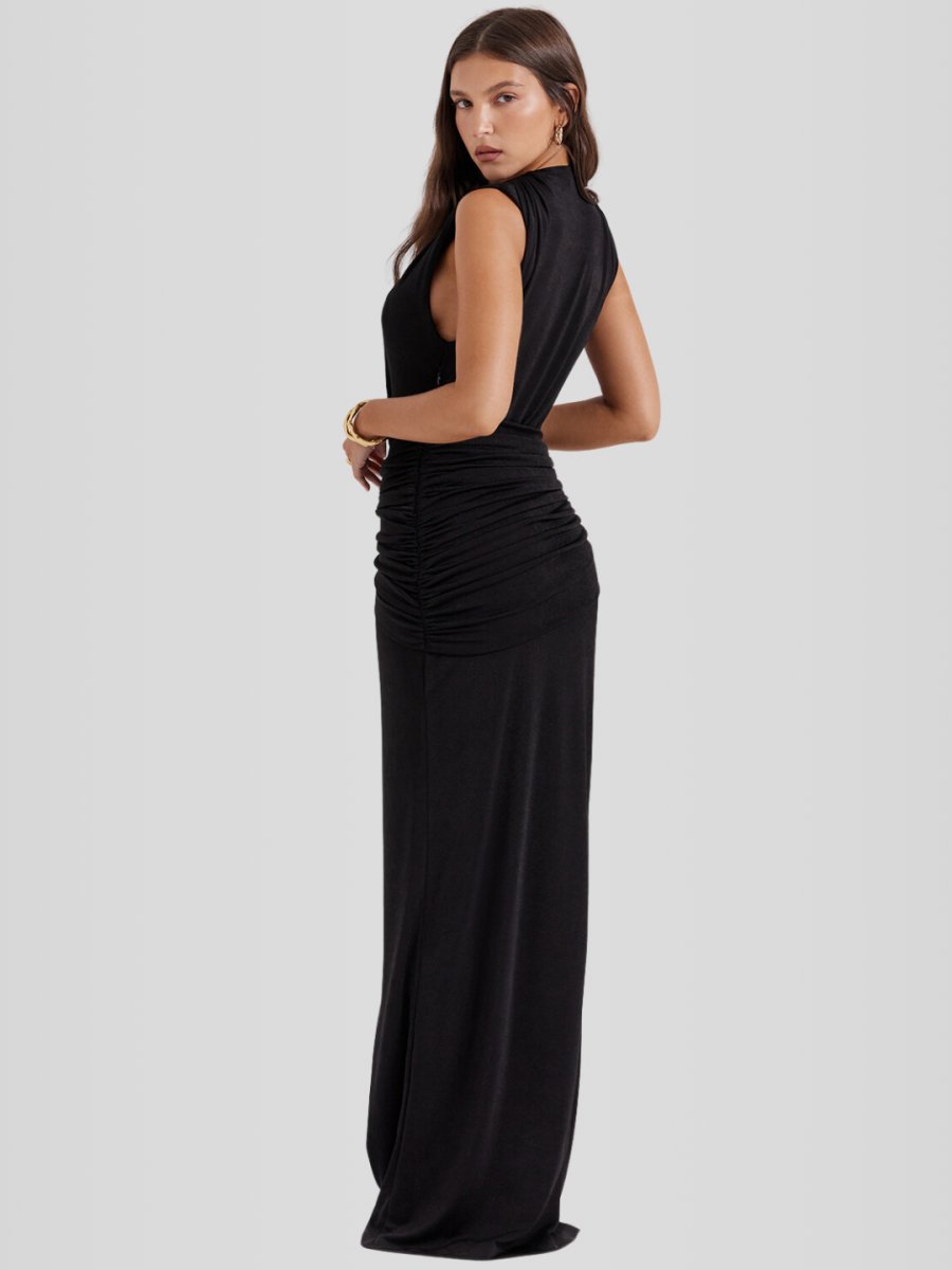 Modelo usando vestido largo negro con escote profundo en V, sin mangas, drapeado frontal y abertura alta en la pierna. Diseño ajustado, elegante y sofisticado.
